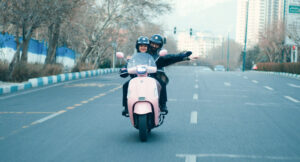 Mann und Frau sitzen af einem pinken Mofa und fahren durch eine Stadt.